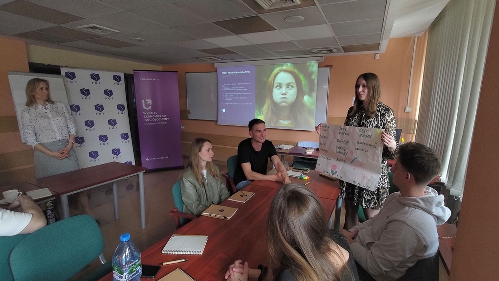 szkolenie pt. Autoprezentacja i radzenie sobie ze stresem - Polska Grupa Farmaceutyczna - 14.04.2025 - studenci siedzący przy stołach i Prowadząca szkolenie patrzą na plakat z tekstem trzymany w dłoniach przez studentkę