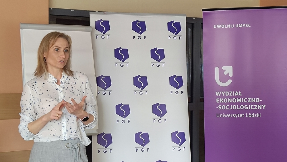 szkolenie pt. Autoprezentacja i radzenie sobie ze stresem - Polska Grupa Farmaceutyczna - 14.04.2025 - Prowadząca szkolenie stoi na tle rollupów PGF i Wydziału Ekonomiczno-Socjologicznego UŁ