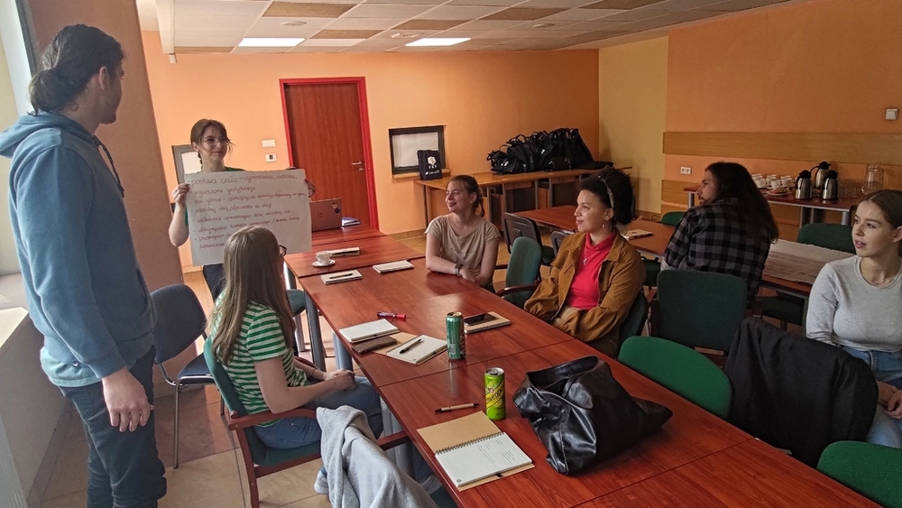 szkolenie pt. Autoprezentacja i radzenie sobie ze stresem - Polska Grupa Farmaceutyczna - 14.04.2025 - studenci siedzący przy stołach i patrzą na plakat z tekstem trzymany w dłoniach przez studentkę