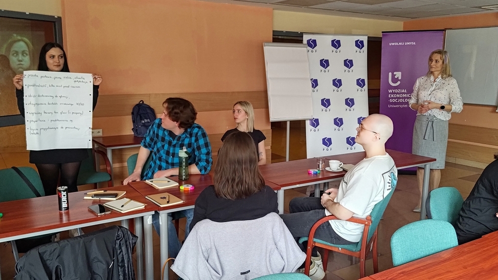 szkolenie pt. Autoprezentacja i radzenie sobie ze stresem - Polska Grupa Farmaceutyczna - 14.04.2025 - studenci siedzący przy stołach i Prowadząca szkolenie patrzą na plakat z tekstem trzymany w dłoniach przez studentkę